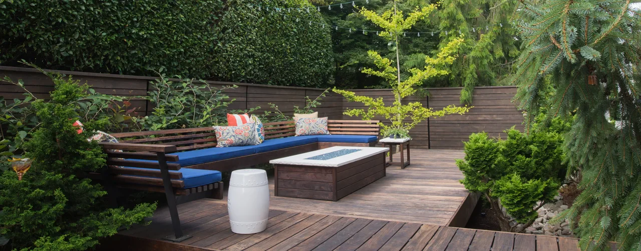 A cozy outdoor lounge with a wooden deck, comfortable seating, a fire pit, and lush greenery, creating a relaxing ambiance
