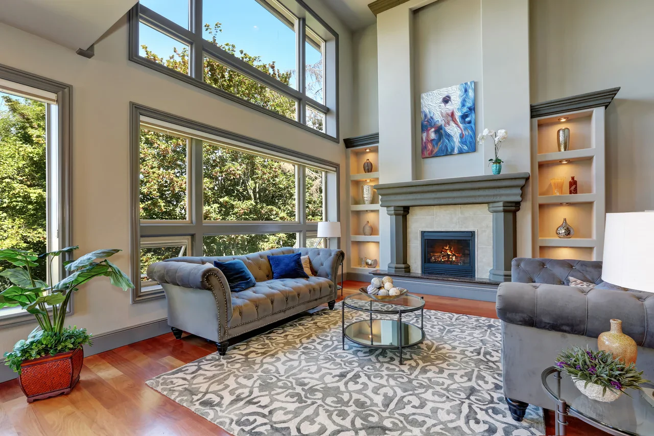 Spacious modern living room featuring a gray tufted sofa, glass coffee table, fireplace, and large windows with natural light.