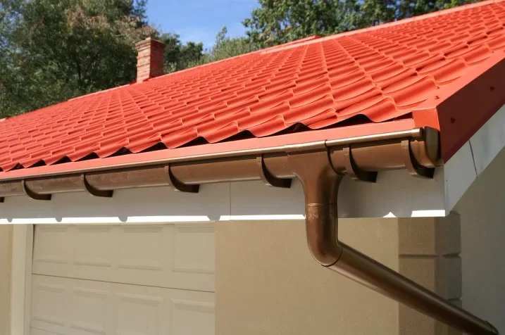 Close-up of a red-tiled roof with brown gutters, showcasing a modern residential design against a backdrop of green trees.