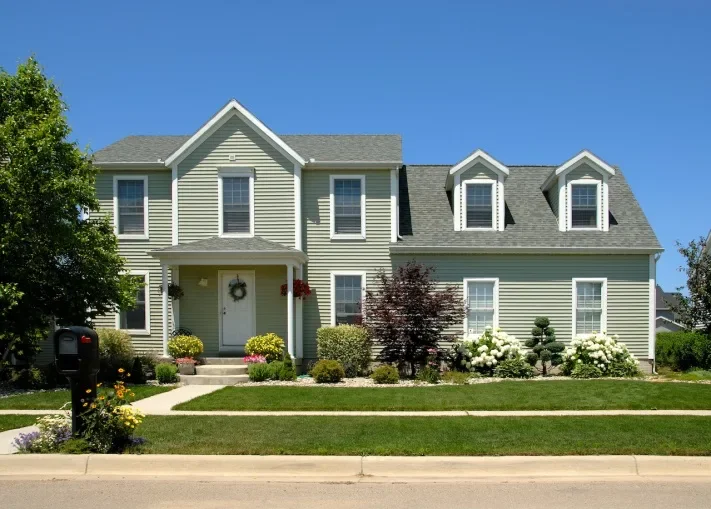 A charming two-story light green house with a gray roof, surrounded by lush gardens and blue sky. A mailbox is visible in front.
