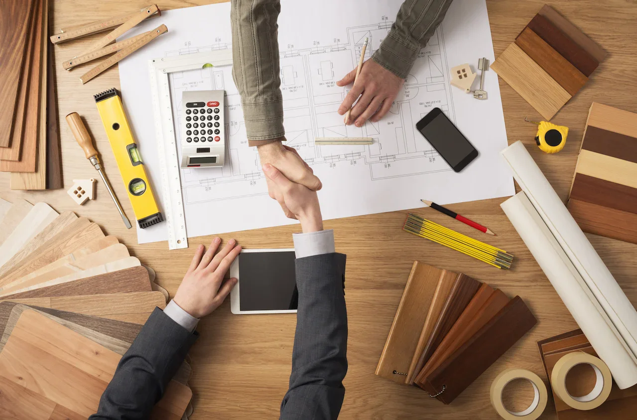 Two people shake hands over blueprints and wood samples, surrounded by tools like a calculator, level, and measuring tape on a wooden table.