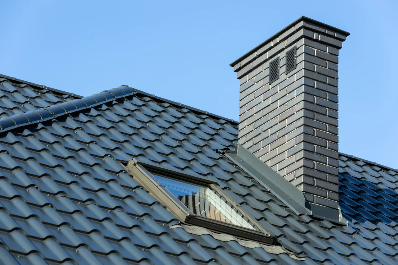 Close-up of a modern roof featuring textured blue tiles, a skylight, and a brick chimney against a clear blue sky.